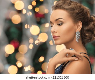 People, Holidays And Glamour Concept - Beautiful Woman In Evening Dress Wearing Ring And Earrings Over Christmas Lights Background