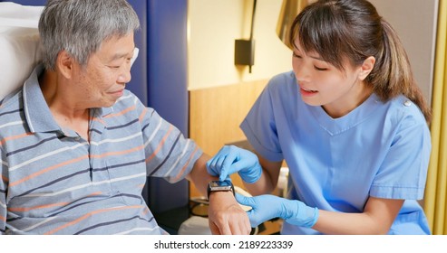 People Home Healthcare Concept - Asian Female Nursing Assistant Is Explaining Health Wearable Smart Watch To Senior Man Who Is Lying On Bed