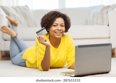 People, Internet Bank, Online Shopping, Technology And E-money Concept - Happy African American Young Woman Lying On Floor With Laptop Computer And Credit Card At Home