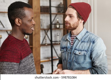 People And Interracial Friendship Concept. Candid Shot Of Two Stylish Male Best Friends Of Different Races Standing Opposite Each Other And Talking Against Modern Restautant Interior Background