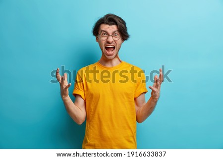 People and irritation concept. Annoyed European man gestures angrily and shouts loudly expresses rage and hate dressed in casual clothes isolated over blue background. Outraged mad guy yells