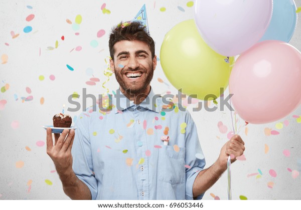人 喜び 楽しみ 幸せのコンセプト 陽気で楽しそうにほほ笑み 絵のポーズをとり カラフルなヘリウム風船とカップケーキを持つリラックスした幸せな誕生日男性 の写真素材 今すぐ編集