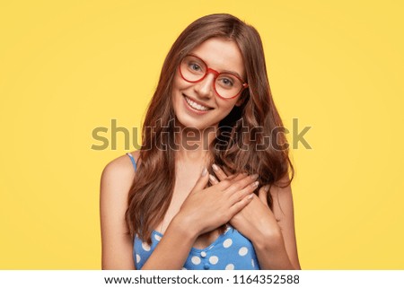 People, kindness and generosity concept. Cheerful young comely woman in spectacles keeps hands on heart, expresses cordialty and friendliness, has charming smile, poses against yellow studio wall