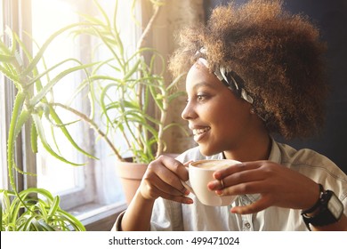 People and leisure concept. Headshot of beautiful African girl wearing bandana and denim shirt, holding cup of hot drink, having tea or coffee, warming up at cafe, sitting near window, looking outside