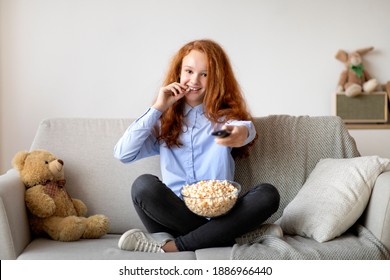 People And Leisure Concept. Portrait of smiling young teenage girl watching tv, eating popcorn from glass bowl in her living room, holding remote control, resting and relaxing sitting on the couch