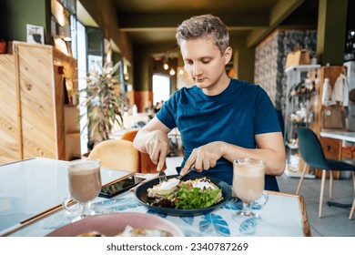 People, leisure and food concept. Young Caucasian man eating some healthy delicious meal with fork and knife at restaurant. Male traveler having breakfast or lunch in cozy cafe. Selective focus - Powered by Shutterstock