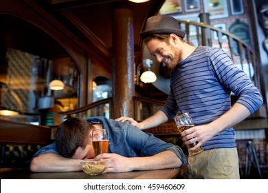 people, leisure, friendship and party concept - man with beer waking his drunk friend sleeping on table at bar or pub