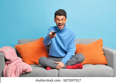 People leisure pastime concept. Overjoyed unshaven adult man sits in lotus poses on sofa holds remote control and watches funny show on television feels entertained enjoys lazy weekend at home