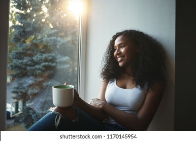 People, leisure, rest, relaxation and lifestyle concept. Picture of fashionable girl with wavy hair smiling happily, having nice time at home, sitting on windowsill and drinking herbal tea - Powered by Shutterstock