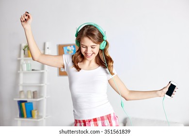 People, Leisure And Technology Concept - Happy Woman Or Teenage Girl In Headphones Listening To Music From Smartphone And Dancing On Bed At Home