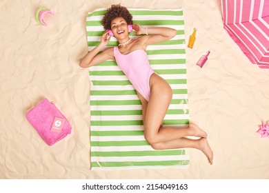 People leisure and vacation concept. Cheerful curly haired beautiful woman dressed in swimwear enjoys favorite playlist while lying in sun at beach has lazy day unforgettable summer holidays - Powered by Shutterstock