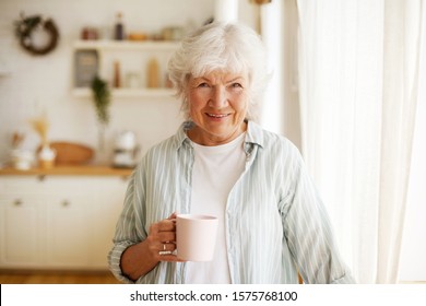 People, lifestyle, age and retirement. Waist up image of cheerful happy European female pensioner relaxing at home, having herbal tea, smiling broadly at camera, posing against kitchen background