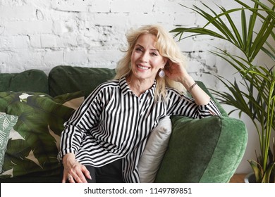 People, Lifestyle And Aging Concept. Portrait Of Friendly Looking Positive Sixty Year Old Caucasian Female With Blonde Colored Hair Relaxing At Home, Sitting Comfortably On Green Couch And Smiling