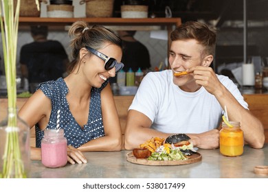 People And Lifestyle. Beautiful Couple Having Fun During Lunch At Coffee Shop, Sitting At Table With Drinks And Food. Attractive Man Eating French Fries And Flirting With His Laughing Girlfriend