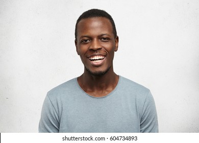 People And Lifestyle Concept. Portrait Of Attractive Young African Male In Good Mood, Wearing Casual Long Sleeve Shirt Smiling Cheerfully Showing His Perfect White Teeth, Happy With Positive News