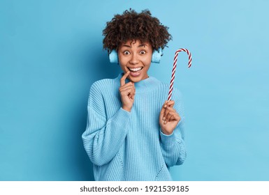 People lifestyle electronic devices concept. Positive good looking Afro American woman listens muisc track via wireless headphones holds delicious sweet candy cane wears sweater isolated on blue