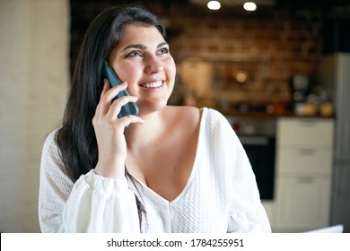 People, lifestyle, electronic gadgets and communication concept. Headshot of beautiful young female with black hair and curvy body smiling happily, receiving good news while talking on mobile phone