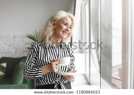 People, lifestyle, food and beverage concept. Portrait of fashionable elegant sixty year old lady wearing stylish striped blouse, having relaxed happy look, enjoying hot drink by the window