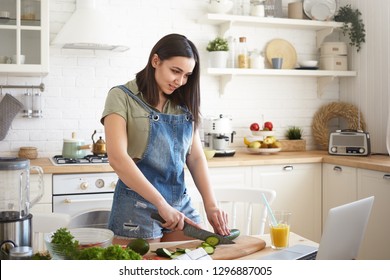 People, Lifestyle, Healthy Eating, Cooking And Dieting Concept. Positive Young Brunette Woman Standing At Kitchen Table, Chopping Green Cucumber For Dinner, Watching Culinary TV Show Online On Laptop