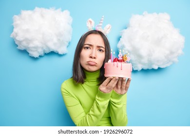 People Lifestyle Holiday Concept. Unhappy Gloomy Asian Woman Holds Tasty Cake Celebrates Birthday Alone Sad About Getting Older Wears Green Poloneck Poses Indoor Against Blue Background With Clouds