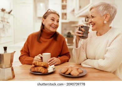 People, lifestyle and leisure concept. Portrait of stylish senior women in cozy sweaters having breakfast together at cafe, sitting at wooden table, sharing news, drinking coffee with pastries