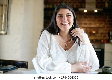People, Lifestyle, Leisure And Modern Technology Concept. Attractive Joyful Young Female Freelancer With Chubby Cheeks Smiling Broadly Listening To Music Via Earphones While Working From Home