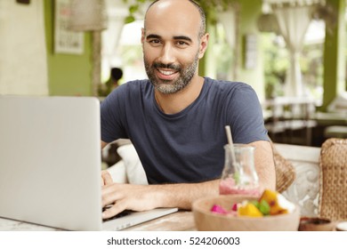 People, lifestyle and modern technology. Outdoor portrait of happy handsome man sitting at cafe table with glass of smoothie and food during breakfast, keeping hands on keyboard of his generic laptop - Powered by Shutterstock