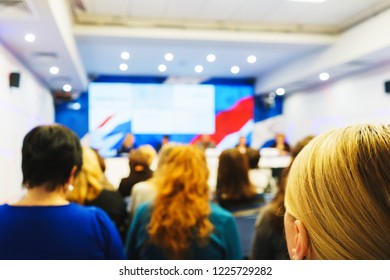 People Listen To The Speaker In The Audience During A Press Conference, Event, Lecture Or Presentation From Behind. Blurred Image Of Participants Of The Conference In The Lecture Hall. Reportage.