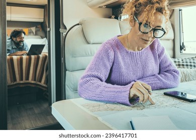 People man and woman in van life day inside camper van. Woman using phone gps map to plan next destination and man working on computer in background. Couple in travel lifestyle with vehicle on caravan - Shutterstock ID 2396417701