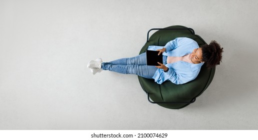 People And Modern Technology. Above top view of young smiling black lady holding tablet sitting on bean bag in living room, watching video. Cheerful lady surfing internet, free copy space, panorama
