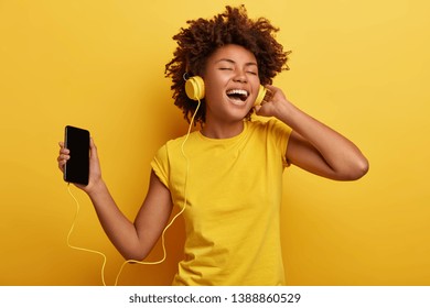 People, Music, Emotions Concept. Delighted Carefree Female With Afro Hairstyle Dances In Rhythm Of Melody, Closes Eyes Listens Loud Song In Headphones, Holds Smart Phone Has Fun. Yellow Color Prevails