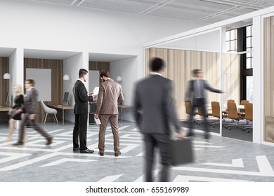 People near office cubicles in an office with white and wooden walls. There are blank vertical pictures in each of them, a desk with a computer, a chair and shelves.
