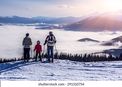 People Observing Mountain Scenery.
Family Of Three People Stays In Front Of Scenic Landscape. These Are Skiers, They Dressed In Winter Sport Jackets And Have Skies Attached.