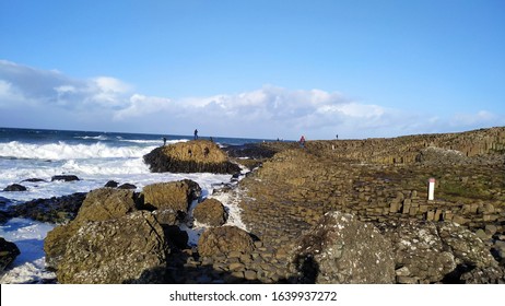 People On The Beach At The Bottom Of The Mountain Slope Formed By Natural Stone And Stacked In A Hexagonal Shape Called The Giant's Causeway In Northern Ireland With The Blue And Rainy Sky