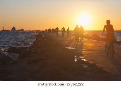 People On Bikes Ride Across The Causeway To The Lighthouse In The Baltic Sea With Ships At Sunset