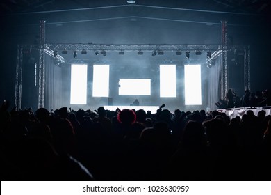 People on the dance floor during a concert in a hangar. Five white led wall ideal to insert text or logos. Laser and lights illuminate the party. Wide view. Dj on the stage