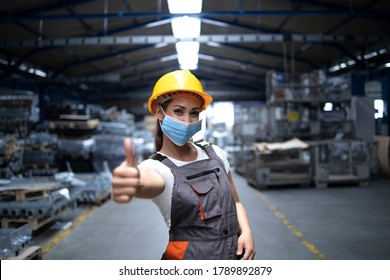 People On Work Obliged To Wear Face Protection Mask. Work Woman Standing In Factory Hall And Showing Thumbs Up While Wearing Hygienic Mask As Prevention Against Corona Virus.