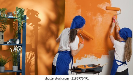 People Painting Walls With Orange Color And Roller Brush, Using Tools And Equipment To Paint Home Interior Decor. Mother And Young Child Having Fun Redecorating Apartment Room. Tripod Shot.