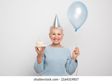 People, Party, Aging And Maturity Concept. Beauitful Middle Aged Female Wearing Conical Hat Celebrating Birthday, Posing Isolated With Cupcake With Candle And Blue Balloon In Her Hands. Happy Time.