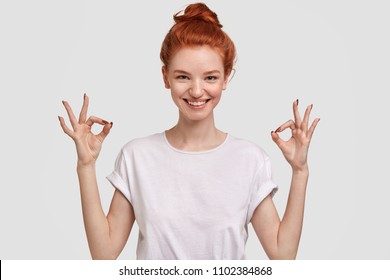 People, peace, body language and meditation concept. Cheerful freckled young female with glad expression, shows ok sign with both hands, dressd in casual white t shirt, demonstrates approval. - Powered by Shutterstock