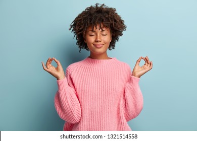 People, Peace And Meditation Concept. Calm Black Young Woman Practices Yoga Indoor, Shows Okay Sign With Both Hands, Demonstrates Approval, Wears Pink Clothing, Isolated Over Blue Background