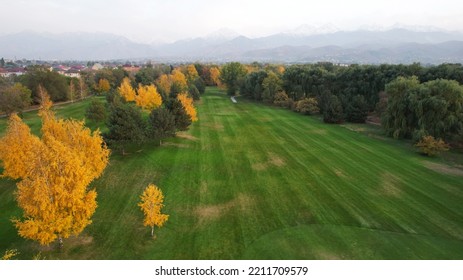 People Play Golf On A Green Field In Autumn. Yellow-red Leaves On Some Trees. Places For Holes. There Is A Golf Car. Houses And Mountains Are Visible In The Distance In A Haze. Sunset. Wedding