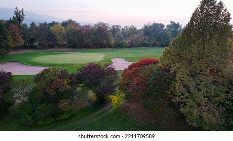 People Play Golf On A Green Field In Autumn. Yellow-red Leaves On Some Trees. Places For Holes. There Is A Golf Car. Houses And Mountains Are Visible In The Distance In A Haze. Sunset. Wedding