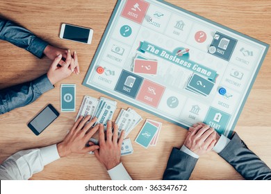 People Playing A Business Board Game On A Wooden Table, One Player Is Holding A Lot Of Money And Winning, Hands Top View