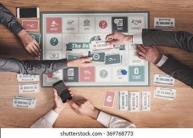 People Playing A Business Board Game On A Wooden Table, One Player Is Paying With Banknotes And Another Is Holding A Card, Hands Close Up, Top View