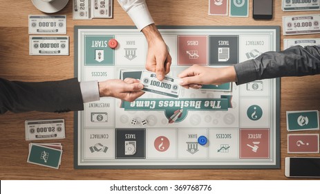 People Playing A Business Board Game On A Wooden Table Challenging Each Other And Holding A Banknote