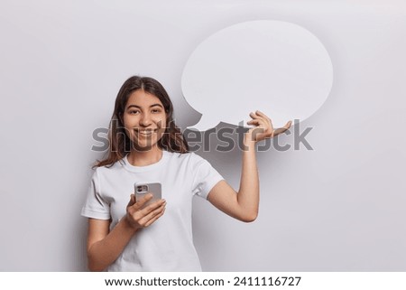 People and positive emotions. Indoor photo of happy smiling Iranian girl standing in centre isolated on white background wearing t shirt holding smartphone and speech bubble with space for promotion