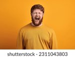 People and power concept. Serious foxy man with determined expression, keeps clenched fists, looks with stern at camera, ready to fight, dressed in dark blue shirt, stands against white wall.