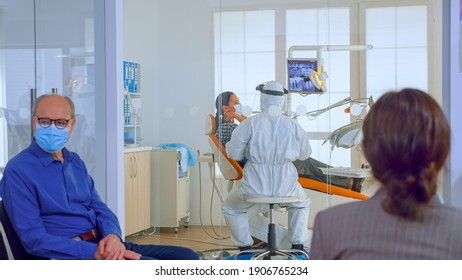 People With Protection Masks Discussing In Reception Area Waiting Doctor In Dental Clinic While Stomatologist Working In Background Wearing Ppe Suit. Concept Of New Normal Dentist Visit