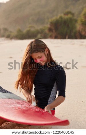 People, recreateion, lifestyle concept. Busy female surfer waxes surface of board with special wax, prepares for surfing competitions, dressed in black wetsuit, models against sandy beach background